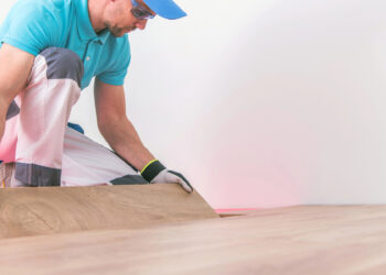 A worker in a blue shirt and protective gloves is installing a wooden floor, bending over to carefully place or adjust a plank.