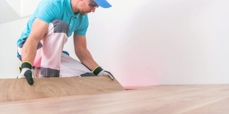 A worker in a blue shirt and protective gloves is installing a wooden floor, bending over to carefully place or adjust a plank.